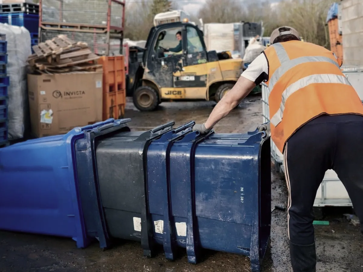 Worker in a high-visibility vest moving a row of wheelie bins in a recycling yard, with a forklift behind